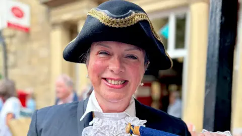 Jaz Oldham A woman in a tricorn hat wearing a white shirt and blue jacket. She is smiling at the camera