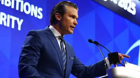Pete Hegseth wearing a dark navy suit speaks at a lectern while pointing his finger at the audience. In the background is a blue screen displaying the Shangri-la Dialogue's name and topic of Hegseth's speech.