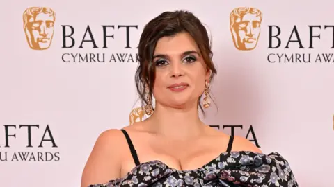 Getty Images A woman with brown hair, dark eye makeup, gold earrings, and a blue and dark floral dress poses for a photo on the Bafta Cymru red carpet, with Bafta images on a white wall behind her.