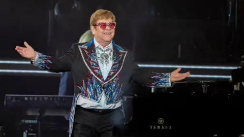 Elton John is standing on stage in a mostly dark jacket. He's wearing sunglasses with red lenses.