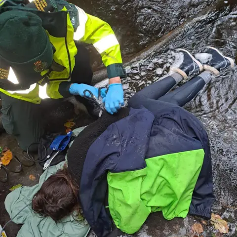 Leah Matthews A woman lying on cold wet ground, covered in coats and blankets. Crouched beside her is someone in high viz uniform, wearing a green beanie and blue medical gloves. 