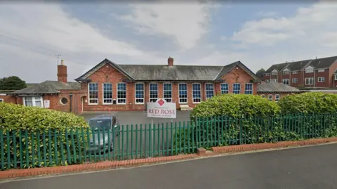 Green gates line a path. Bushes are behind them and a car park. A sign in the middle of the fence reads 'Welcome to Red Rose Primary School'. A red brick building sits behind the car park with large, long white windows. 