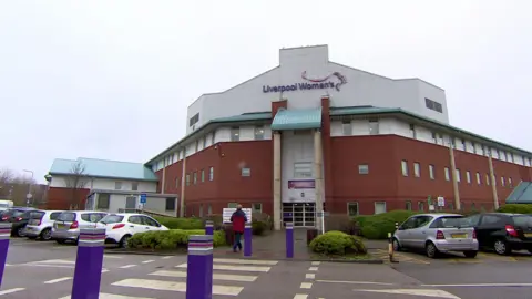 Exterior shot of Liverpool Women's Hospital, the main building is orange, the roof is white. Liverpool Women's is written on the front in purple. Purple bollards lead up to the entrance.