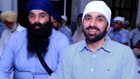 Guppreet Singh Johal Gurpreet dan Jagtar Singh Johal duduk di sebuah ruangan dengan pria Sikh lainnya. Gurprret mengenakan sorban biru kerajaan dan kemeja biru muda dan memiliki jenggot yang panjang dan gelap. Jagtar tersenyum dan mengenakan sorban putih dan kemeja berwarna biru dan putih.