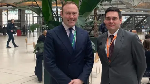 Blake Stephenson Blake Stephenson is in a navy double-breasted suit, wearing a tie and pink shirt. Beside him is a man with a grey suit and spotted tie. They appear to be standing in Portcullis House at Westminster.