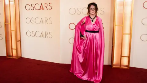 Getty Images Maggie is bright pink long satin dress, black belt, black necklace & little veil hat.
Posing behind the word Oscars.