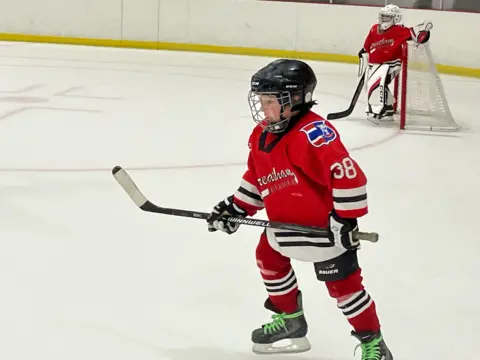 Guy's and St Thomas Trust Rufus Quinn, wearing a red Streatham ice hockey jersey with the number 38, skates on the ice during a game, holding his hockey stick. In the background, the team's goalie stands in front of the net.