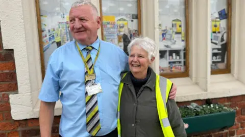 Ray Badrock, gekleed in een blauw overhemd met korte mouwen en een geel en grijs gestreepte stropdas, met een Merseyrail-sleutelkoord en naamplaatje, poseert met zijn arm om de schouders van Margaret Walton