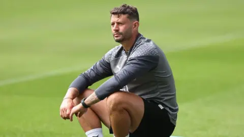 A man in grey and black football training kit sitting on a pitch.
