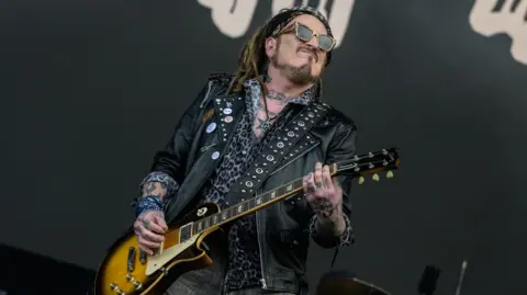 Getty Images A man stands on a stage holding a guitar. He has dreadlocked hair and is wearing a bandana, sunglasses, a black leather jacket with metal studs, and a leopard-print shirt. He has tattoos on his hands, arms and neck