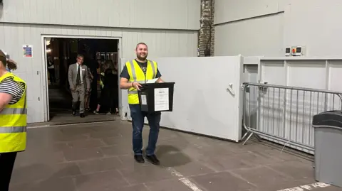 Simon Dedman/BBC A man in high viz carrying a black plastic ballot box into a building.