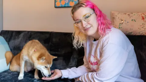 WOODGREEN PETS CHARITY Ciara Pollen is smiling at the camera while Fern the dog eats treats from her outstretched hand. Ciara has blonde and pink wavy, shoulder-length hair and is wearing a pale pink sweatshirt. She and the dog are sitting on a sofa in a living room.