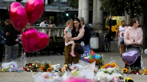 Reuters A woman holding a child looks at flowers and tributes left for victims of Monday's attack in Southport