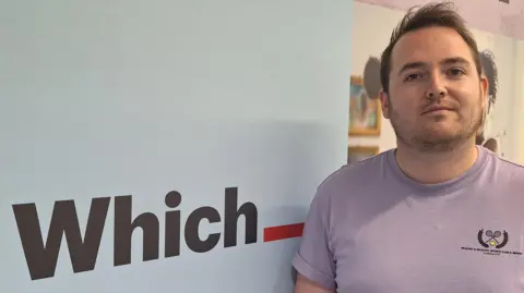Which? Rob Lilley-Jones wears a purple t-shirt and is standing in front of a Which" sign. He is white, with brown hair and stubble.