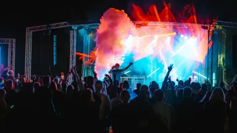 Supplied A dark image taken from behind a large audience showing a band performing on stage. There are colourful lights on the stage and smoke. 