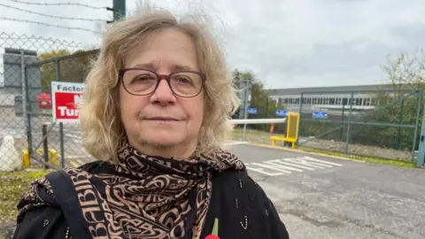 Chris Willmore, a middle-aged woman with glasses and fair hair wearing a black and orange scarf. She is standing in front of the factory site.