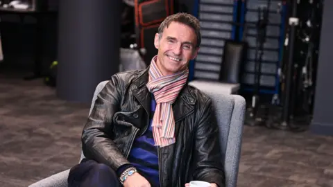 Marti is sitting on a grey lounge chair and smiling at the camera. He is wearing a black leather jacket, blue and black top and a stripy scarf. He has short brown hair.