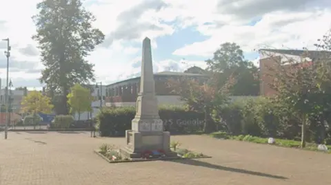 Google A small courtyard area with a war memorial in its centre. It is stone and tall with a square base that gets thinner towards the top. There are red poppy wreaths at its base, and small bits of greenery planted in a border around it.