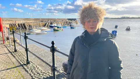A woman in a coat and cossack-style hat stands in front of a harbour with some boats bobbing around. There is a harbour curving around on the left to her. The sky is blue, but features dark clouds.