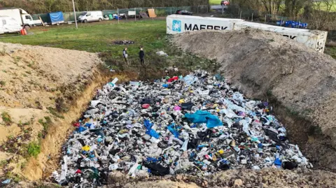Picture shows a large amount of rubbish dumped in a three-sided pit. Two people can be seen in the background, along with several vehicles and containers.