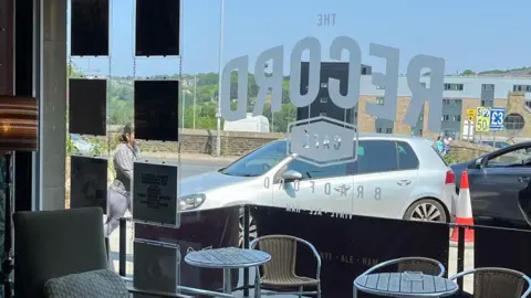 Keith Wildman The inside of a bar looking out through a plate glass, floor to ceiling window which shows a silver-coloured car parked close to tables outside. 