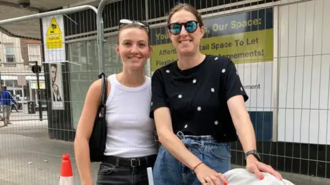 Alex Pope/BBC Alex Reader, wearing a white top and black jeans, with Lauren Steels, wearing sunglasses, a dark top with flowers on and blue jeans, standing outside Bedford's former Debenhams store, which has fencing up outside 