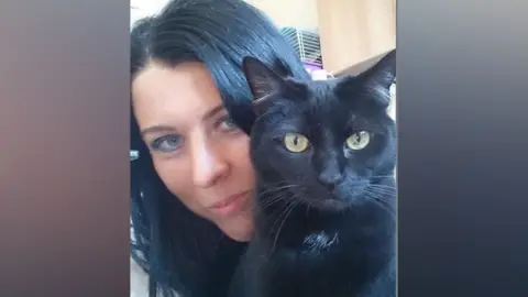 Mandy Hobbis A woman with black hair takes a selfie with a black cat. The cat has a small patch of white fur on its chest. 