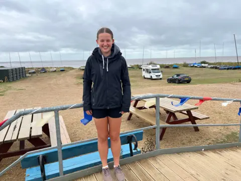 Amelia Eaton leaning on the railings at Snettisham Beach Sailing Club. She is wearing black shorts and a dark blue fleece. Behind her are a row of boats and the sea