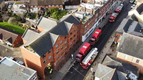 Maldon Fire Station Drone image, taken at an angle, of three fire engines and two red lorries on an otherwise empty High Street. Several fire fighters and police officers are standing around. In the centre of the image a red-brick building has its gable end of bricks missing. Rubble can be seen on a flat roof next door and there is an alleyway between the two buildings. Houses and gardens can be seen behind the buildings.