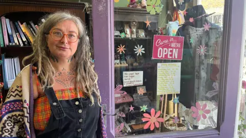 Rosie Coates has long hair and is wearing red framed glasses, black dungarees over a yellow and orange checked top and a cream and purple knitted cardigan. She is stood in the doorway of her shop which is painted purple with hand painted flowers on the glass.