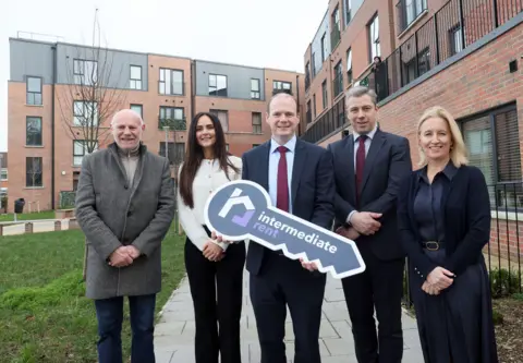 Department for Communities Minister Gordon Lyons holds a large cardboard key which says "intermediate rent". He's accompanied two men and two women 