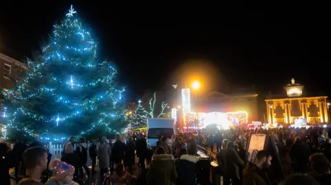 A night time Christmas light switch on in King's Lynn, showing a light Christmas tree, light up buildings, people standing bout, a white van, and street lights.