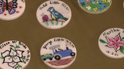 6 white badges with various designs sit on a brown table cloth. The bottom is a pink plant on the right side and the date is 15/03/21. In the middle is a blue tractor with name and date in the badge, George Little, 16/03/20. Above it is a pink ball of wool with a blue tight and knitting needles, it says Violet Little, 17/01/21. The bottom is a white flowers and Elene McBrid named, 19/12/21. The top left has a picture of a purple and orange butterfly. The top is of a garden filled with different types of flowers on the right side. 