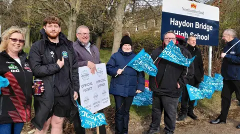 LDRS Seven teachers waving blue flags outside a blue and white school sign which reads Haydon Bridge High School. One man is holding a sign which says Official Picke while another points to a blue sticker on their jacket. They are all smiling. 