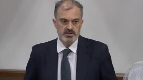 Parliament TV A man with blading grey hair and a short beard. He is stood up and is wearing a dark navy suit, white shirt and dark grey tie. 