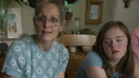 A woman with tied up grey hair wearing light-coloured round glasses and a blue jumper sits next to a teenage girl with long brown hair and blue T shirt.