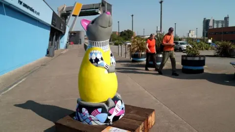A rat sculpture painted with a yellow jumper and displayed on a wooden board outside The Deep