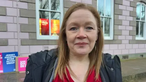 A woman with ginger hair looks into the lens of the camera. She is wearing a red jacket under a black coat. Behind her, signs in favour of assisted dying rest against a pink wall. 