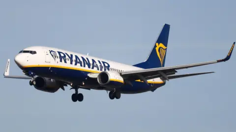 A blue, white and yellow Ryanair plane with the company's logo on it shown in mid-flight against a blue sky. 
