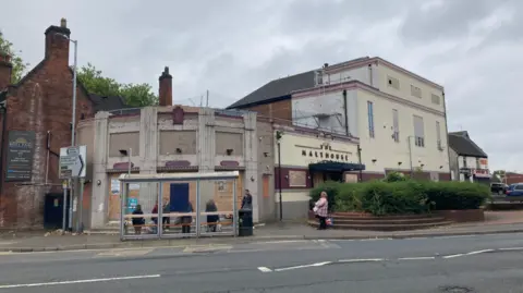 The view across a road to a derelict building with boarded up windows, with the words The Malthouse visible on the wall. The building varies in height, and is curved on the left hand corner where it meets the road