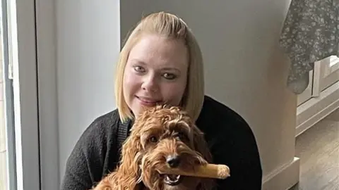 Charlotte Alderson, who has blonde-bobbed hair, wearing a dark grey or black cardigan. She is holding a sandy-coloured dog, which has a bone in its mouth.