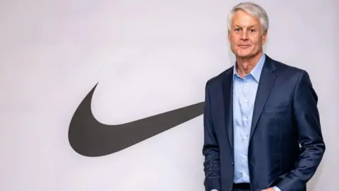 Former Nike chief executive John Donahoe poses for a photograph, smiling in front of the company's swooshing tick logo. He has white-grey hair and wears a navy suit.