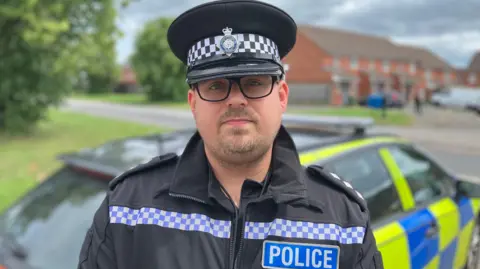 Sam Read/BBC Ch Insp Nathan Shepherd looks at the camera, wearing a police cap and black jacket with the word POLICE on the front. Behind him is a neon yellow and blue police car.