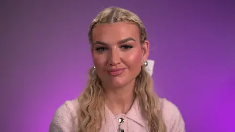 Sacha Jones looks at the camera in front of a pink background.