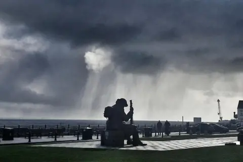 BBC Weather Watchers / MackemIan A statue of a man in military gear with his head bowed is illuminated in silhouette. Rain clouds are overhead.