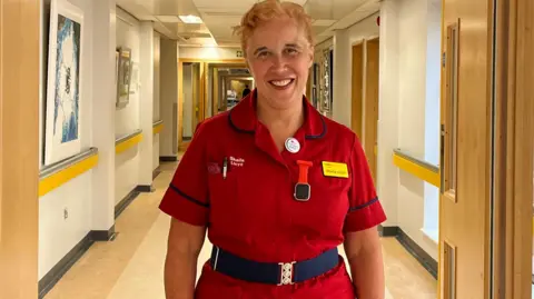 Sheila Lloyd, a woman with blonde hair in a bun and a bright smile, she is wearing a red nursing dress and is standing in the hospital corridor.