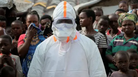 Reuters A health worker in personal protective equipment (PPE) stands near displaced people waiting for the burial of suspected Ebola victims at the Kigonze displaced persons camp, one month after an outbreak was declared, in Bunia, eastern Democratic Republic of Congo, June 18, 2026.