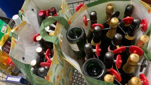 Two large vinyl shopping bags containing about 30 bottles of alcohol (mostly wine or champagne) seen from a bird's eye view in a shopping trolley. To their left are a handful of other items such as chocolates and laundry detergent. 