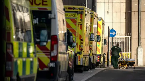 PA Media A row of ambulances queuing to get into a hospital. A paramedic in the distance is pushing an empty stretcher.