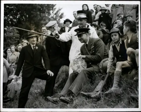 Getty Images A black-and-white photograph showing a crowd gathered on a grassy hillside, where an official holds a long stick while another person seated nearby holds a round cheese, watched closely by onlookers including children, during a Cooper’s Hill cheese rolling event.